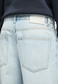Jean en denim bleu clair avec poches arrière visibles et passants de ceinture, porté avec un haut foncé rentrée à la taille.