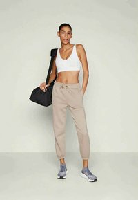 Γυναίκα που φοράει λευκό αθλητικό σουτιέν, μπεζ φόρμες, γκρι αθλητικά παπούτσια, κρατώντας μια μαύρη τσάντα γυμναστηρίου στον ώμο, στέκεται μπροστά σε απλή ανοιχτόχρωμη φόντο.