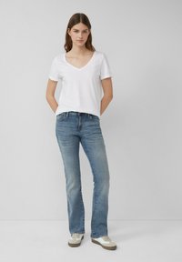 Witte v-hals t-shirt met opgerolde mouwen, lichtblauwe rechte jeans en beige sneakers met witte accenten. Model staat tegen een grijze achtergrond.