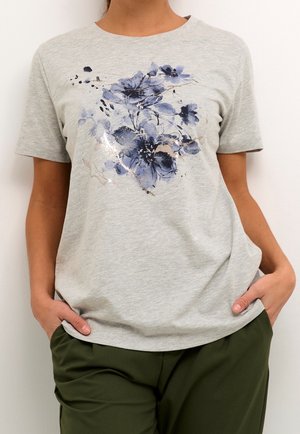Kvinde iført lysegrå T-shirt med blåt blomsterprint og olivengrønne bukser, hænderne i lommerne, foran en ensfarvet hvid baggrund.