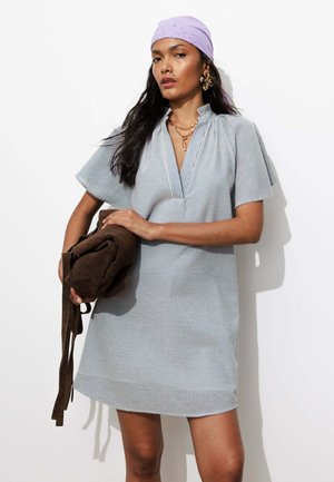 Femme portant une robe à manches courtes rayée et un foulard lavande, tenant un sac en daim marron, avec des boucles d’oreilles en or et des colliers superposés.