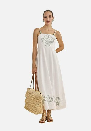 Mujer con vestido blanco sin mangas con bordado floral, sosteniendo un bolso tote beige con flecos, llevando sandalias doradas y pendientes verdes.