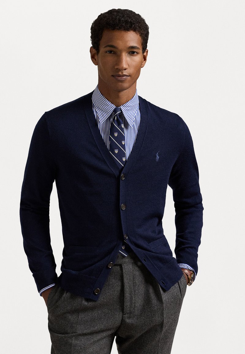 Navy knit cardigan with a V-neck, button front, and two pockets. Worn over a blue and white striped dress shirt and patterned tie.