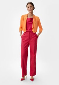 Jasnopomarańczowy krótki kardigan, różowa satynowa bluzka i czerwone spodnie z wysokim stanem. Modelka stoi z rękami w kieszeniach, nosząc czerwone szpilki.