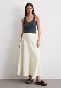 Débardeur en vert foncé, associé à une jupe longue couleur crème en tissu léger ; des sandales noires texturées complètent le look.