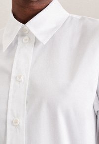 Vit skjorta med knappar framtill, som har en klassisk krage, runda knappar och en slät tygtextur. Enkelt mönster med ett rent och fräscht utseende.