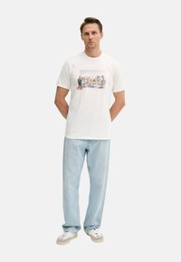 Camiseta de algodón blanca con un estampado gráfico que presenta "GUESS" y cactus. Combinada con jeans de mezclilla azul claro y zapatos blancos con detalles en gris.