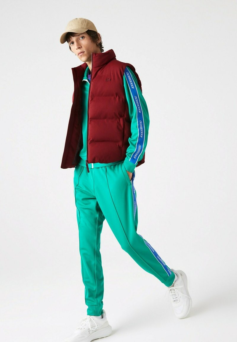 Bordeauxfarbene Steppweste über einem grünen Trainingsanzug, der blaue Seitenstreifen und weiße Sportschuhe hat. Eine schlichte Mütze rundet den sportlichen Look ab.