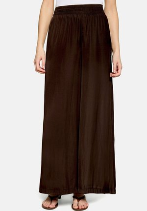 Femme portant un pantalon large marron foncé avec une ceinture élastique et des sandales à bout ouvert, se tenant devant un fond uni.