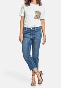 Žena v bílém tričku s květinovou kapsou, modrých zkrácených džínech s roztrhanými lemy a béžových sandálech na podpatku, stojí zkříženě.