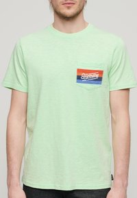 Camiseta de manga corta verde claro hecha de una tela suave, que cuenta con un pequeño bolsillo en el pecho con un diseño de logo multicolor de Superdry.