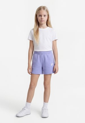 Jeune fille debout portant un t-shirt blanc, un short Nike violet, des chaussettes blanches et des baskets blanches sur un fond blanc uni.