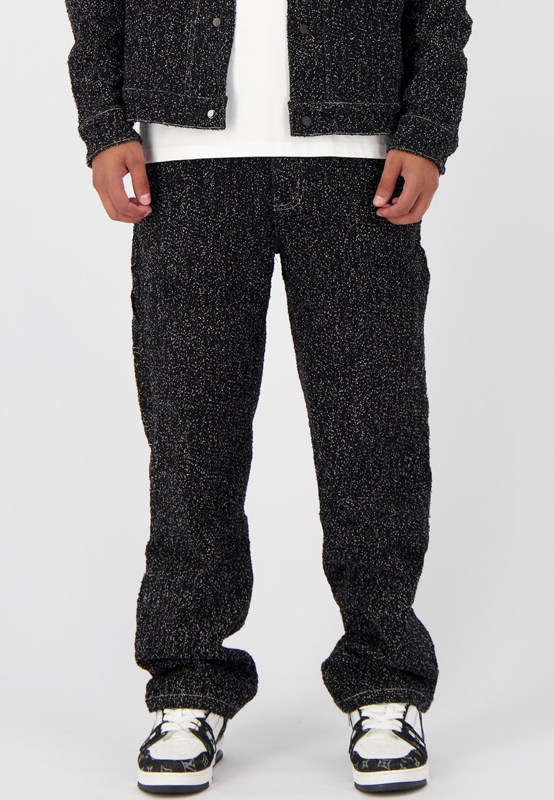 Zwart-witte gespikkelde broek met een ontspannen pasvorm. De stof heeft een gestructureerde afwerking. G gedragen met een witte t-shirt en gemusterde sneakers.