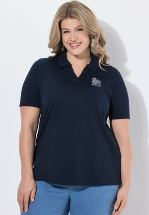 Smilende kvinde med blondt hår iført en navy polo-shirt med floralt "BLOOM" logo og lyseblå jeans, står foran en hvid baggrund.
