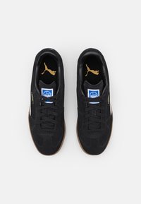 Svarta mockaskor med en vit rand, svarta snören och en sula av gummi. Har en guldfärgad logotyp på tungan och hälen.