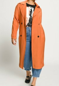 Pomarańczowy płaszcz trencz z wyciętym kołnierzem, podwójnym zapięciem z przodu i teksturowanym materiałem; noszony na czarnej koszulce z nadrukiem i niebieskich dżinsach.
