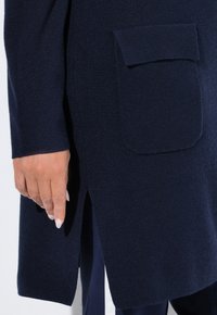 Detailní záběr ruky vedle námořnické modré pletené kardiganu s klopovým kapsou a bočním rozparkem, nošeného přes odpovídající kalhoty s bílými pruhy.