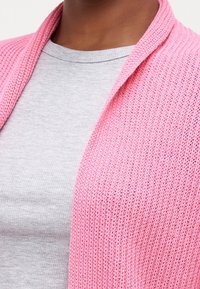 Svetle růžový pletený kardigan s texturovaným vzorem, nošený přes šedé tričko s kulatým výstřihem; má otevřený přední díl a šálový límec.