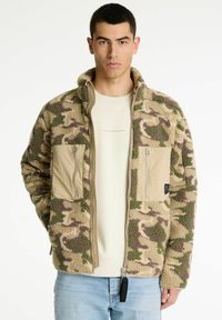 Fleecejas in tan met een camouflagepatroon in groen en bruin. Voorzien van twee voorzakken met rits en een hoge kraag. Ritsluiting aan de voorkant.