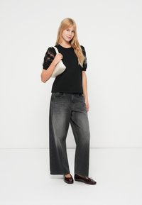 Μαύρο μπλουζάκι με μανίκια από δαντέλα και φουσκωτά, συνδυασμένο με παντελόνι φαρδύ σκουρόγκρι και μπορντό φλατ παπούτσια. Το μοντέλο κρατά μια μικρή κρεμ τσάντα χειρός.