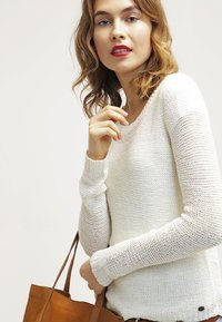 Pull en tricot blanc avec de longues manches texturées, dotée d'un col rond. Le mannequin tient un sac fourre-tout en cuir brun.