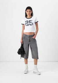 White cropped t-shirt with navy trim and printed number "25," paired with gray wide-leg denim shorts, white sneakers, and a black patterned bag.