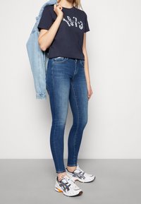  camiseta de algodón azul marino con diseño gráfico blanco, combinada con jeans skinny de mezclilla azul. Superpuesta con una chaqueta de mezclilla azul claro y zapatillas blancas.