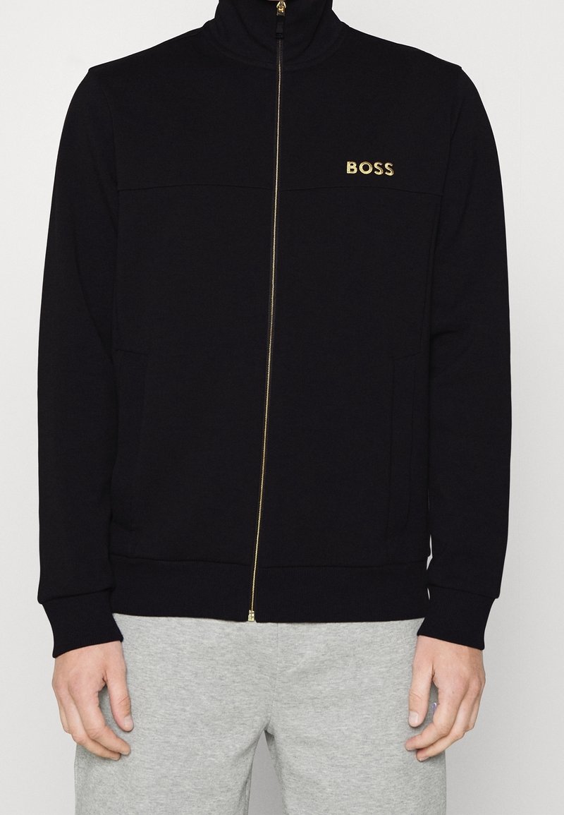 Veste noire à fermeture éclair dorée avec logo "BOSS" sur la poitrine gauche, portée avec un pantalon gris clair, montrant le torse et les mains.