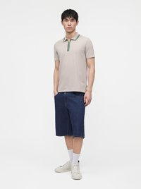 Jeune homme portant un polo beige à manches courtes, un short en jean bleu foncé jusqu'aux genoux, des chaussettes blanches et des baskets blanches, debout devant un fond uni.