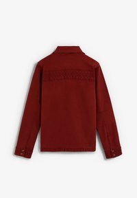 Chaqueta de pana roja con un diseño de yugo texturizado, mangas largas, puños abotonados y corte recto. Sin herrajes visibles en la vista trasera.