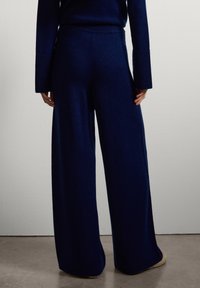 Personne portant un pantalon en maille large bleu marine et un pull assorti, se tenant sur un sol en béton contre un mur blanc.