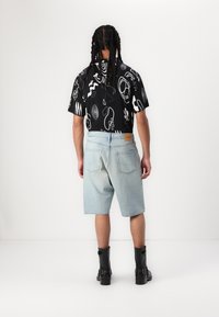 Shorts de mezclilla cortos en azul claro, con dos bolsillos traseros. Camiseta negra con un patrón abstracto en blanco. El modelo lleva botas negras hasta el tobillo.