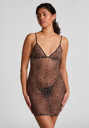 Frau trägt ein durchsichtiges Slipkleid mit Leopardenmuster und dünnen schwarzen Trägern, posiert vor einem schlichten hellgrauen Hintergrund.