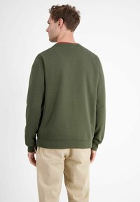 Olijfgroene sweater met een ronde halslijn, geribbelde manchetten en zoom, en rode accenten bij de kraag, gedragen over beige broeken.