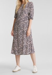 Femme portant une robe midi fleurie bleu marine avec des manches trois-quarts et des baskets blanches, debout devant un fond uni.