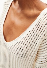 Pull en tricot avec un col en V profond, fabriqué dans un tissu texturé de couleur crème présentant des rayures verticales et des mailles ouvertes.