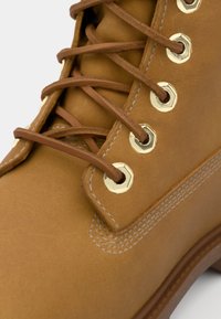 Bota de cuero marrón con ojales de metal en tono dorado, cordones marrones y costuras blancas. Textura suave y diseño redondeado.