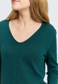 Turquoise gebreide trui met een diepe V-hals en lange mouwen, met een zachte textuur en naadloos ontwerp, waarbij de aansluitende pasvorm wordt benadrukt.