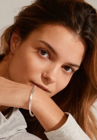 Silver metallic bracelet with a textured, zigzag pattern worn on a wrist. The background features soft, neutral tones.