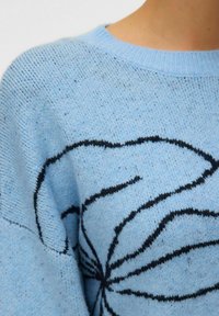 Svjetloplava džemper od pletene tkanine s teksturiranim završetkom. Ima crni cvjetni uzorak na prsima i okrugli izrez.