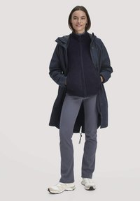 Marine waterdichte parka over een donkere fleece vest met rits, gecombineerd met lichtgrijze strakke broek en witte hardloopschoenen. Glad textuur van de stoffen is duidelijk zichtbaar.