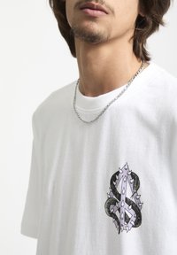 T-shirt en coton blanc présentant un graphique marquant sur la poitrine en violet et noir, représentant un design stylisé avec des pointes et des serpents.