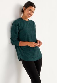 Türkis gestrickter Pullover mit runder Ausschnittlinie, langen Ärmeln und seitlich geschlitztem Saum, kombiniert mit schwarzen Hosen. Strukturiert, lockere Passform.