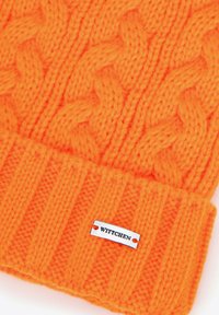 Cappellino arancione lavorato a maglia con motivo a trecce e risvolto a coste. Comprende un'etichetta con logo in metallo cucita sul lato.