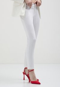 Donna che indossa pantaloni bianchi aderenti e scarpe con tacco alto rosse con cinturino alla caviglia, in piedi su uno sfondo bianco semplice.