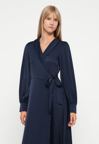 Navy wrap dress in soft fabric with long puff sleeves and a tie at the waist. Features a deep V-neck and smooth texture.