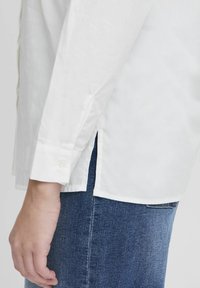Chemise blanche à manches longues en coton, avec des poignets à boutons et des fentes sur les côtés, associée à un jean en denim bleu.