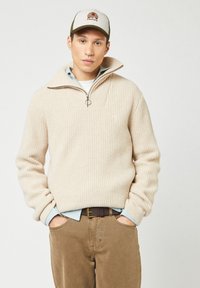 Man in een beige geribde half-zip trui, blauw geblokt overhemd, bruine broek, riem en een beige en groene pet met logo. Handen in de zakken.