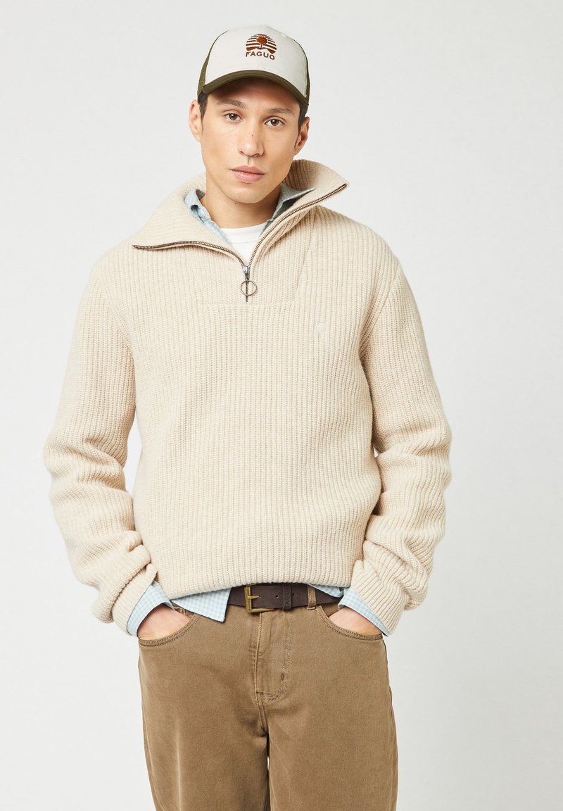Man in een beige geribde half-zip trui, blauw geblokt overhemd, bruine broek, riem en een beige en groene pet met logo. Handen in de zakken.