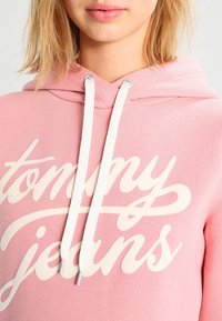 Sudadera rosa con el logotipo "tommy jeans" en letra blanca. Cuenta con una capucha con cordón y una textura suave, mostrando un diseño casual.